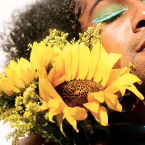 A  non-binary person with Afro hair and bright green eyeshadow holds sunflowers to their face. Text overlay reads: It's a dynamic and diverse ecology